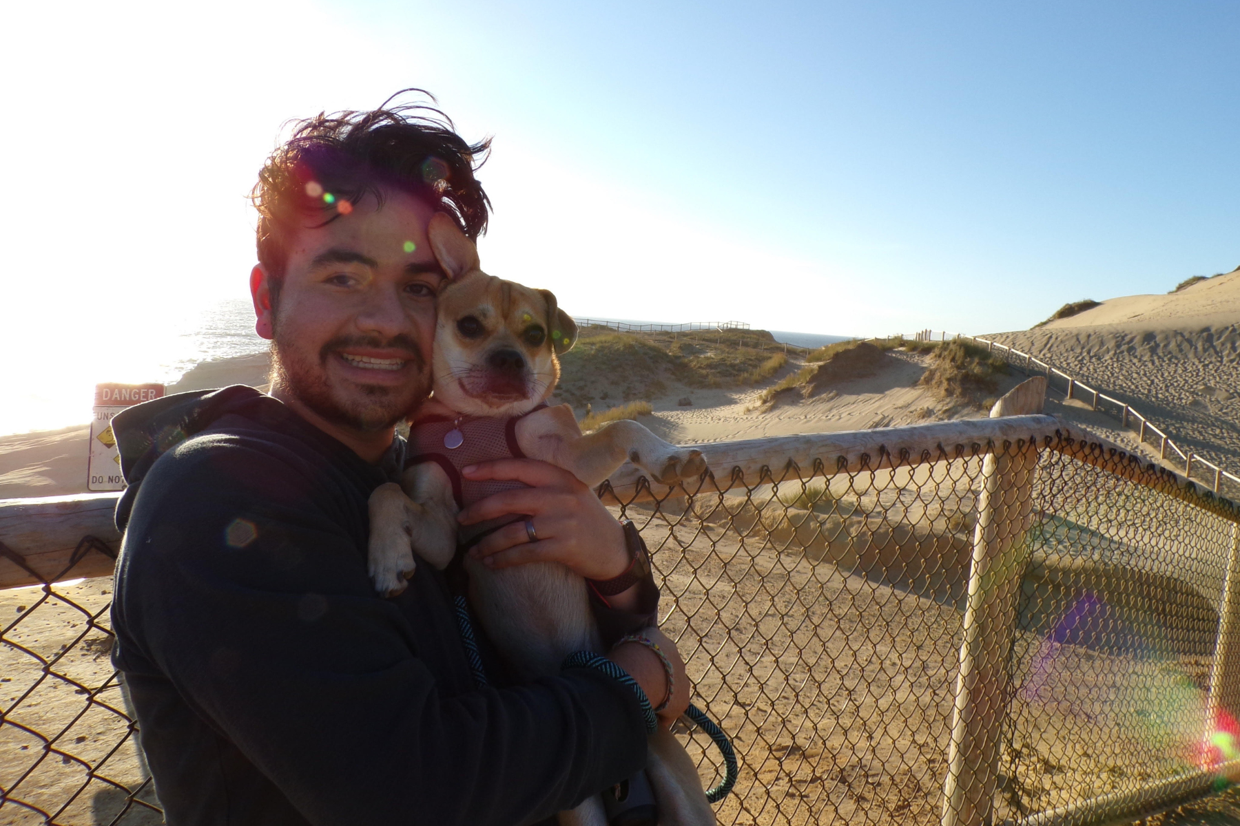 Man standing with a dark hoodie on, holding a small dog. The man is in front of a fence that surrounds sand dunes at a beach.