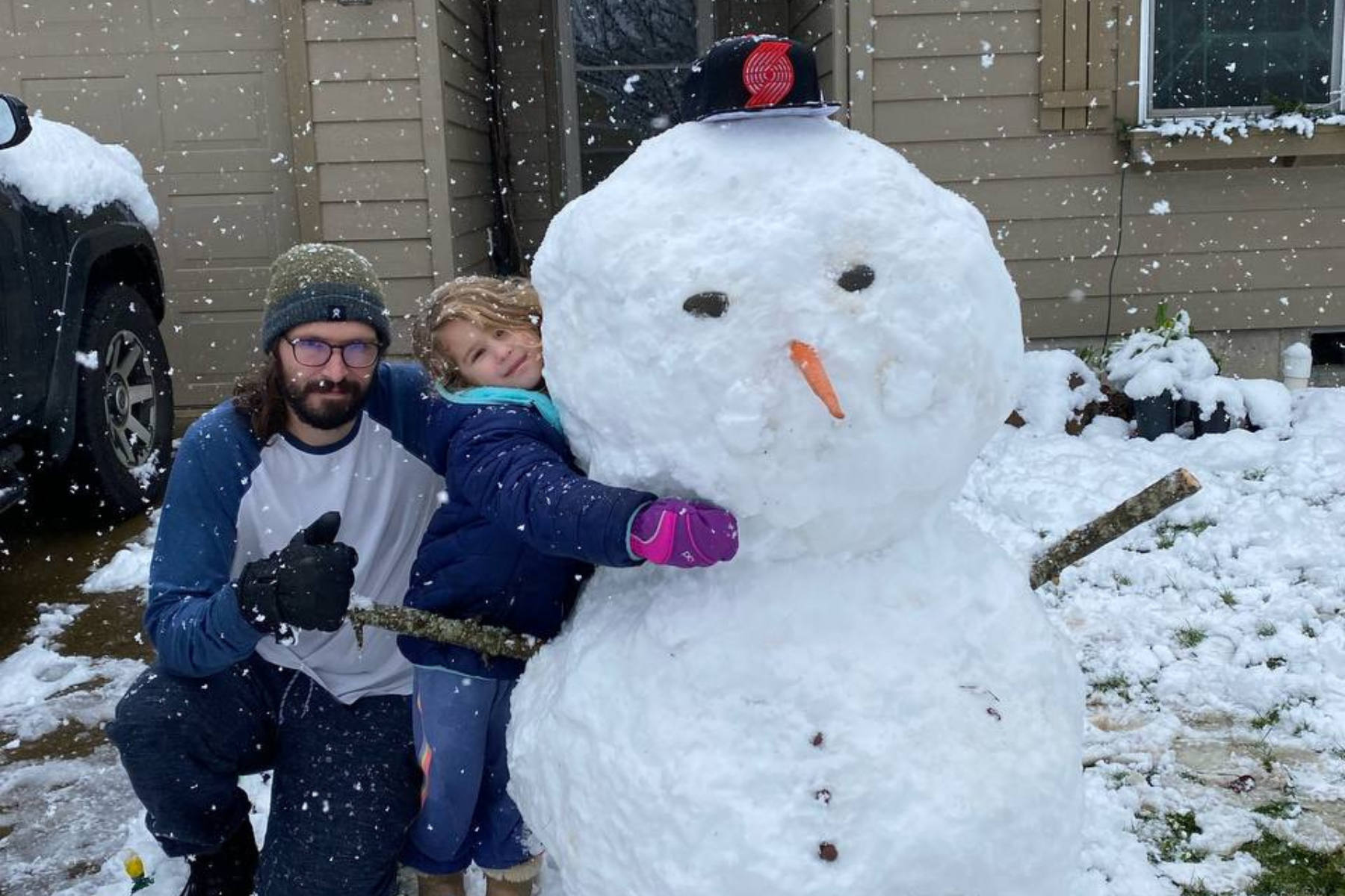 A kneeling man with glasses in a white and blue long sleeve shirt, black gloves, a beanie and sweatpants giving a thumbs up with a blonde child in a blue puffy jacket, jeans, and pink gloves in the driveway of a brown house. The child is hugging a snowman with a trailblazers hat on. It's snowing and there's a layer of snow covering the ground.