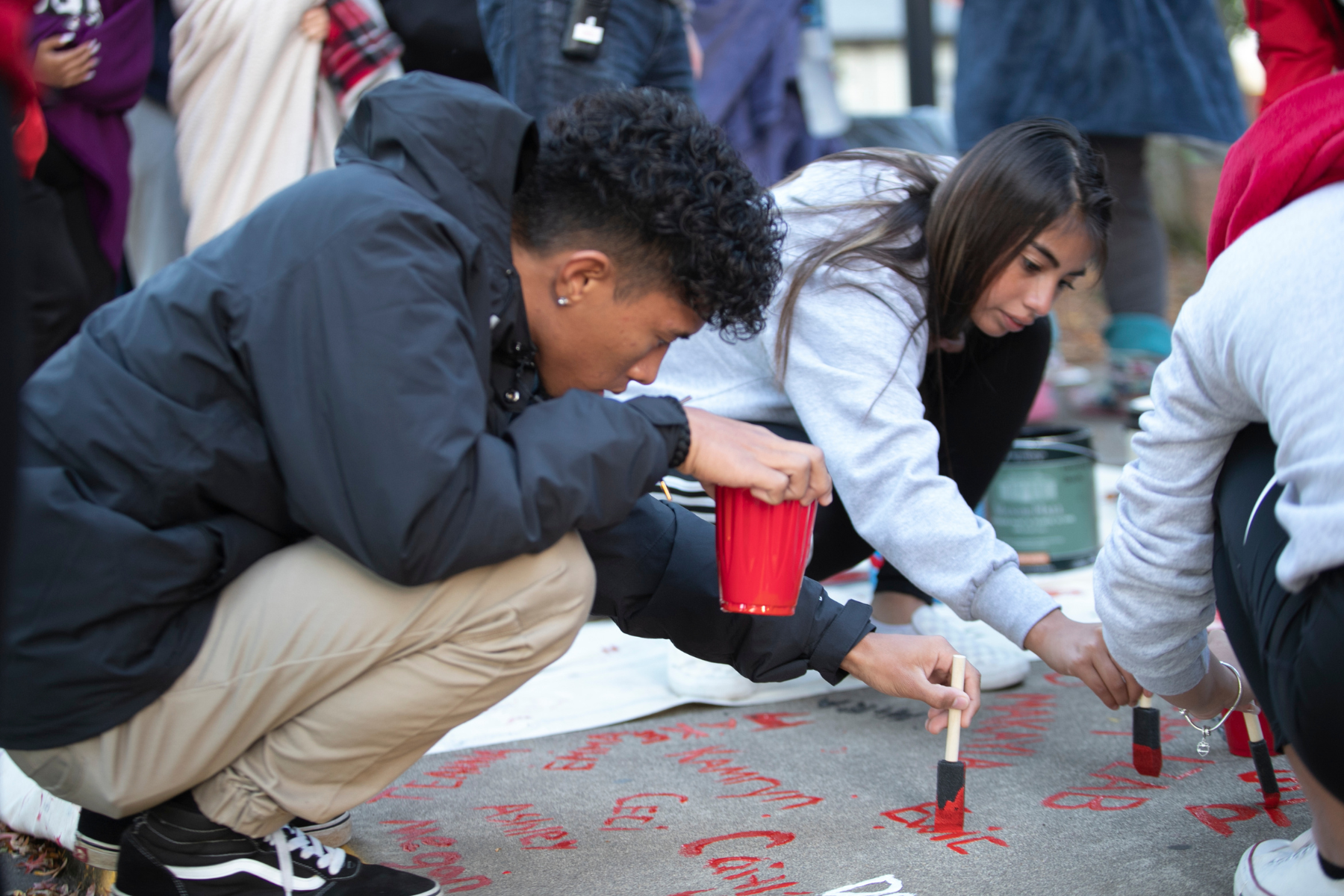 Man squatting near the sidewalk using red paint to paint their name on the sidewalk.