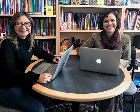 two people with laptops at a table with bookcases in the background