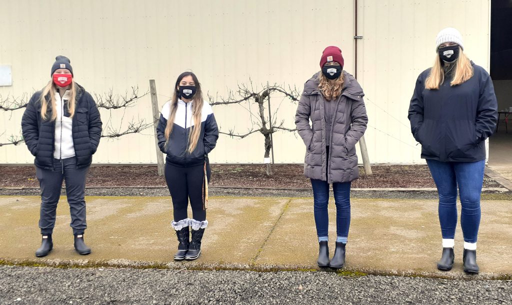 Four students wearing WOU non-surgical masks stand in front of a warehouse.