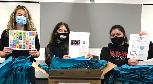 Three students wearing non-surgical masks, holding up informational flyers and standing behind cardboard boxes with supplies.