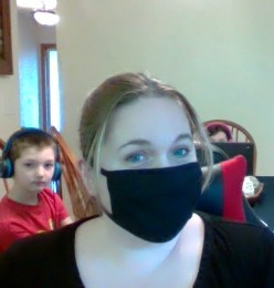 Tracie Wicks wearing a non-surgical mask with her children sitting at a table behind her. Her children are wearing headphones and have laptops. 
