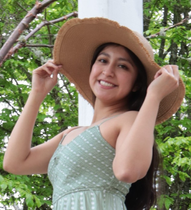 Dana standing in front of foliage with a green dress holding a large sunhat.